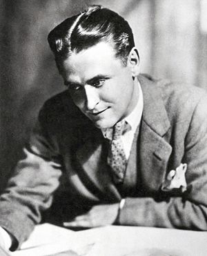 A photograph of F. Scott Fitzgerald by Nickolas Muray. Fitzgerald is bent over a desk and is examining a sheaf of papers. He is wearing a light suit and a polka-dot tie. A white handkerchief is in his breast pocket.