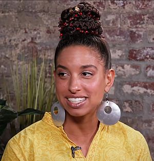 Woman with brown skin looking off to the side wearing silver earrings and yellow shirt with microphone clipped to lapel.