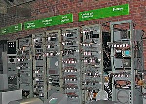 A series of seven tall metal racks filled with electronic equipment standing in front of a brick wall. Signs above each rack describe the functions carried out by the electronics they contain. Three visitors read from information stands to the left of the image