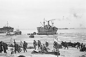 Troops come ashore on one of the Normandy invasion beaches, past the White Ensign of a naval beach party, 7 June 1944. A24012