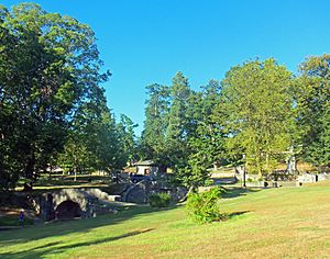 A grassy landscape with trees in the background. In front of them is a small creek with two stone bridges. A tall monument is at right