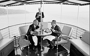 President Johnson has dinner with guest aboard the Sequoia as Secret Service Agent Rufus Youngblood stands in the background, July 15, 1965