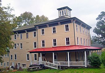 A large yellow wooden building with green trim and a wraparound porch with red metal roof. There is a cupola in the center of the roof and various construction debris on the porch and ground in front.