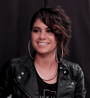 Still image of a woman looking away from the camera and smiling with an undercut hairstyle; wearing large hoop earrings, a black top, a thick necklace, and a leather jacket.