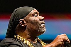 close-up profile of Turbo B wearing a black top and thick gold chain, with a black head covering, holding a microphone in one hand and appearing to perform