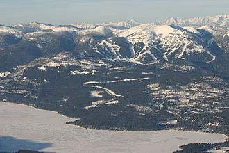 Aerial view of a forested mountainous area, with ski trails.  In the foreground is a partially frozen body of water.