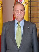 King Carlos in a grey jacket, light blue shirt, yellow tie, standing indoors in front of the Spanish flag