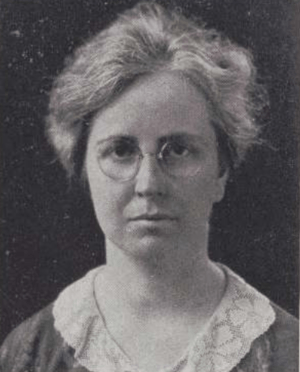 A middle-aged white woman with grey hair, wearing round glasses and a lace collared blouse or dress.