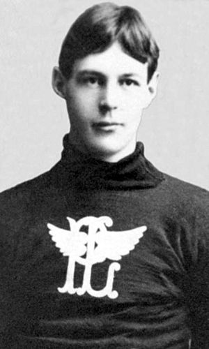 Black and white image of a young man shown from torso up, with arms cropped out of image. Wearing wool sweater with an image, a "P" and "L" overlapping on each other, with wings coming out of the "L" to partially obscure the "P".