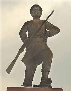A cement statue depicting a man with a moustache holding a rifle in one hand and a stick in another, wearing traditional clothes. He stands on a polished stone pedestal.