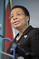 Graça Machel dressed formally in a black jacket, wearing a pearl neckless, speaking on a podium.