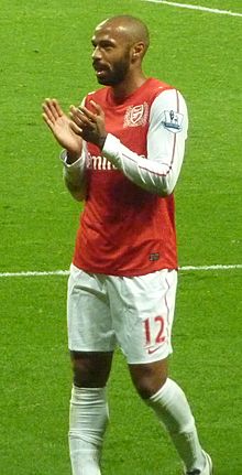 Thierry Henry, wearing a red shirt with white long sleeves and shorts with a number 12 and Nike logo on the left-leg side, applauds.