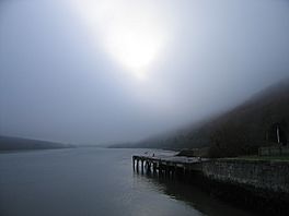 A stone and concrete jetty extends along the shore of a lake between hills under a misty sky