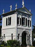 A small, white. roughly square house. It is ornately decorated with statues at the top of the roof gable, worked pillar-like facings, and a gable end pediment with dentil molding. All of the trim is white except the shutters, which are black.