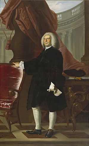 A man in black, 18th-century clothing and stands next to a red chair in front of an imagined Palladian backdrop.