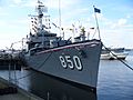 A gray battleship, with the number 850 painted large on its bow, is tied up to a dock.