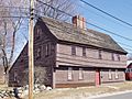 A brown garrison-style saltbox house with a wooden roof and a red door. A rough stone wall separates the street from the yard.