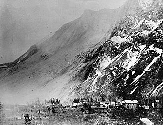 A small town shown at the base of a mountain. The mountain's face stands barren following a large rockslide and a light cloud of dust is visible in the air.