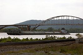 Tugboat drifts down the Arkansas River paralleling a set of railroad tracks while both cross under a long bridge with concrete and steel arches in front of the Ozark Mountains in the background