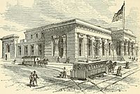 An Egyptian-Revival-style building on a street corner, with doric columns on its façade and a large flag flying atop a mast. Pedestrians are seen navigating two horse-drawn trams, which travel on lines that bisect intersecting streets.
