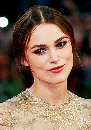 A portrait of Keira Knightley, wearing black tied-up hair, red lipstick, and a gold dress.