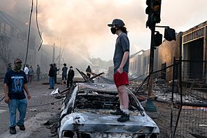 A man stands on a burned out car on Thursday morning as fires burn behind him in the Lake St area of Minneapolis, Minnesota (49945886467).jpg