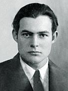 Ernest Hemingway's passport photograph. He is facing the camera with a neutral expession on his face and has short dark hair. He is wearing a dark suit, black tie, and white shirt.