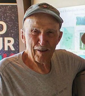 A white man in a grey t-shirt and ballcap is facing and looking into the camera, smiling.