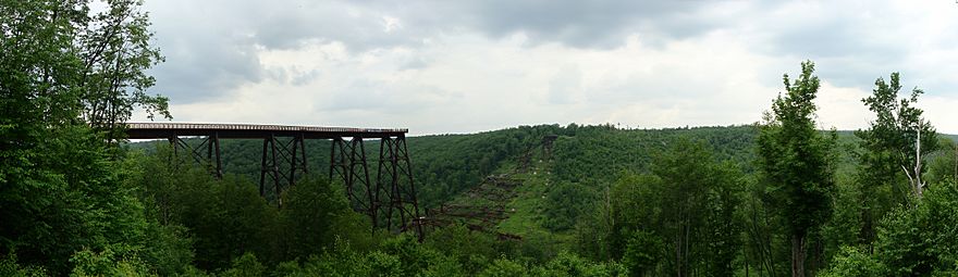 View of the remains of a bridge across a valley. Part of the bridge in foreground and a portion at its far end remain standing, whereas the rest is collapsed and lying on the valley floor.