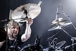 Marco Minneman behind a drum kit, holding a crash cymbal with his left hand after having hit it with a drumstick in his right.