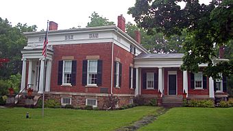 A brick house with a much larger, more decorated, flat-roofed section on the left with four chimneys on top and a smaller, plainer gabled wing on the right with one chimney