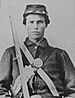 Head and torso of a young white man wearing a full military uniform, including a cap, straps crossing his chest, and a rifle leaning against his right shoulder.