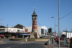 A brown clock tower surrounded by several infrastructure, photographed from across the road.