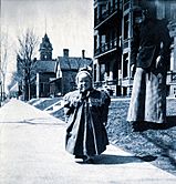 Black-and-white photographic portrait of writer F. Scott Fitzgerald as an unbreeched infant with his mother in Saint Paul, Minnesota. Fitzgerald is standing on a city sidewalk with his mother nearby on the grass. In the distance behind them is a steepled building, likely a church, and several leafless trees.