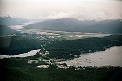 Aerial view shows Auke Bay (including the harbor and Auke Lake) in the foreground.  The Mendenhall Peninsula extends to the right behind the community.  The lower Mendenhall Valley, Juneau International Airport and Douglas Island are in the background.