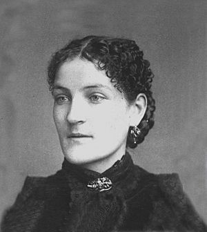 A white woman with dark curly hair, wearing dangling earrings and a high-collared black dress fastened with a broach at the neck.