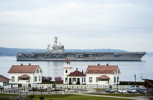 A large aircraft carrier passes behind a white lighthouse and several buildings in a park-like setting.