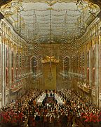A giant hall with gilded white walls with a table with three branches in the middle, surrounded by a crowd. Four people are sitting under a tall golden canopy, the rest next to two longer branches. Most people are standing. The chandeliers and the ceiling is decorated with pink and white flowers and green leaves, and a band in red is playing from two balconies..