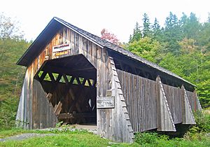 A brown wooden covered bridge with pointed roof. Above its portal are signs saying "Millbrook 1902" and "Pedestrians only". To the right a faded sign says "Safe load 6 tons"