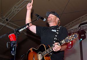 Keith onstage, smiling and holding a guitar behind a microphone while pointing upward