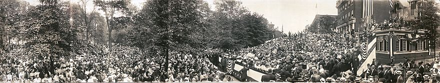 A panorama image showing a large crowd seated in chairs and standing outdoors around a speaker who delivers a speech on a podium in the center. Downtown brick buildings and large campaign posters are in the background.