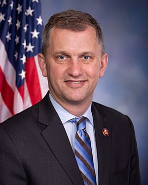 Official portrait of Casten from the 116th Congress. Sitting in front of an American flag, he wears a dark suit, a light blue shirt, and a blue striped tie.