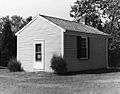 A black and white photograph of a small one-room building. It has a door on one side and a window on another.