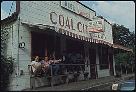 A white building with 'COAL CITY CLUB' in painted in maroon. Some miners sit on benches outside; others "hunker down".