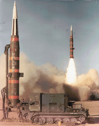 missile erect and prepared for launch while missile is launching in background