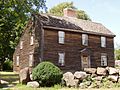 A simple brown saltbox house with a central chimney stands behind a rough stone wall.