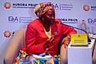 Marguerite Barankitse in a colourful skirt and headdress, and a red jacket. She is speaking on a stage as part of a panel discussion.