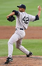Mariano Rivera in a gray baseball uniform and navy blue cap stands on a dirt mound. His right arm is behind him, bent at the elbow and clutching a baseball. The back of his uniform shows the number 42.