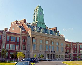 An ornate brick building with a stone face, four stories high with a large green cupola on top, seen from its left with some parked cars in the foreground.