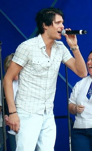 Basshunter, who is dressed in white, sings into microphone while standing in front of people and blue background